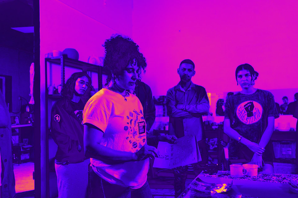 Una persona habla frente a un grupo con playeras de La Testería, en tonos magenta.